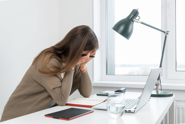 Attractive caucasian woman graphic designer tired of hard work. Sad girl cover face with hands. On table laptop, graphic tablet, glass of water and notepad. Female horrified by the number of tasks. - Photo, Image