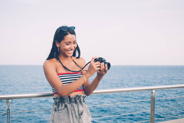 Feminino turista étnico positivo em roupas de verão em pé no calçadão perto do mar com câmera fotográfica vintage e tirar fotos enquanto relaxa durante as férias - Foto, Imagem