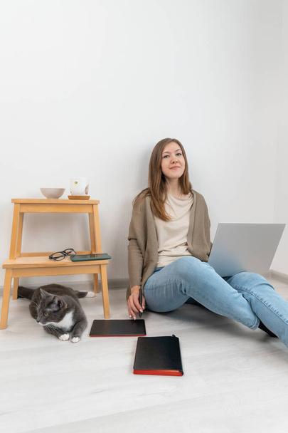 Beautiful woman graphic designer sitting on floor and working on a laptop. Girl in cardigan and blue jeans using digital tablet in work. Female looking into camera. Home office concept. Near grey cat. - Photo, Image