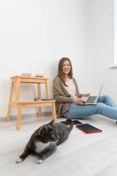Aantrekkelijk brunette meisje met laptop maken een pauze in het werk voor het spelen met kat. Vrouw op de vloer. Nieuw huis zonder meubels. Het concept van thuiskantoor. Selectieve focus op mooie grijze kat.  - Foto, afbeelding