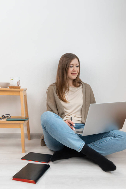 Angenehm lächelnde brünette Frau in grauer Strickjacke und blauer Jeans, die auf dem Boden sitzt, an einem Laptop arbeitet und ins Display schaut. Auf dem Boden schwarzer Notizblock und Grafiktablett. Home Office-Konzept. - Foto, Bild