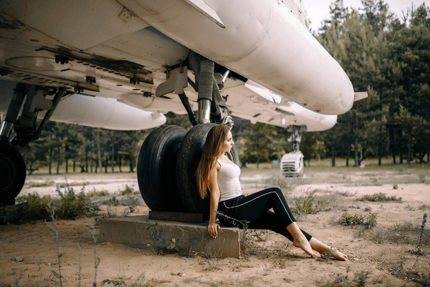 mooi jong brunette meisje staat op de achtergrond van oude militaire vliegtuigen. Meisje in een wit shirt en zwarte broek in de natuur. Militaire uitrusting. Portret van halve lengte. meisje poseren - Foto, afbeelding