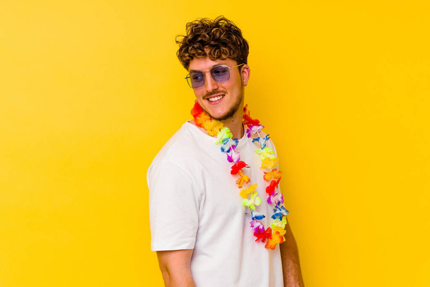 Young caucasian man wearing a Hawaiian party stuff isolated on yellow background looks aside smiling, cheerful and pleasant. - Фото, зображення