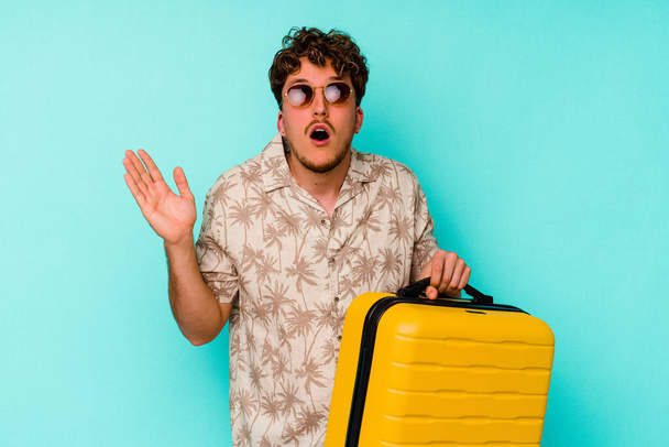 Молодий мандрівник тримає жовту валізу на синьому фоні здивований і шокований
. - Фото, зображення