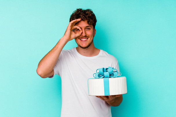 Young caucasian man holding a cake isolated on blue background excited keeping ok gesture on eye. - Фото, изображение