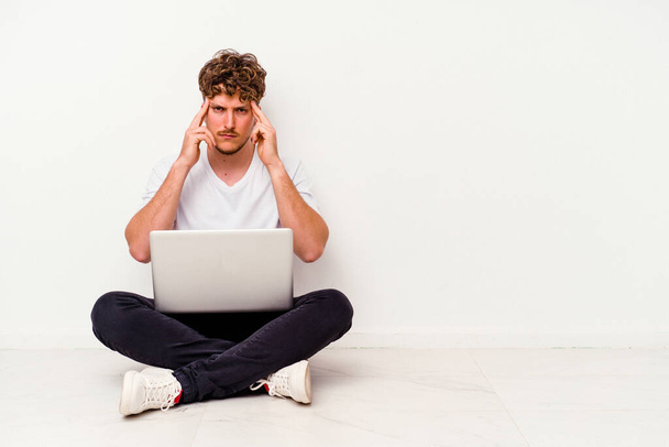 Jonge blanke man die op de vloer zit... met een laptop geïsoleerd op een witte achtergrond... gericht op een taak, met wijsvingers gericht op het hoofd.. - Foto, afbeelding