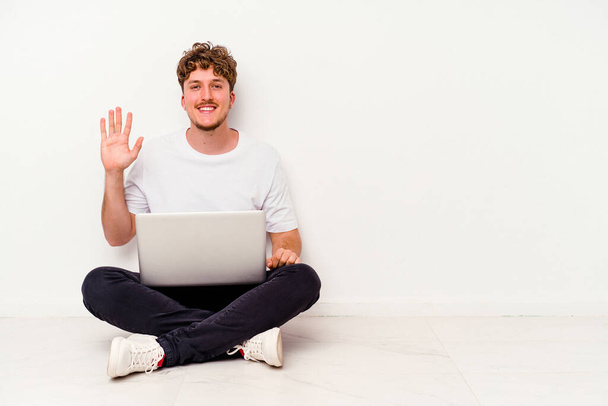 Mladý běloch sedí na podlaze drží na notebooku izolované na bílém pozadí s úsměvem veselá ukazuje číslo pět s prsty. - Fotografie, Obrázek