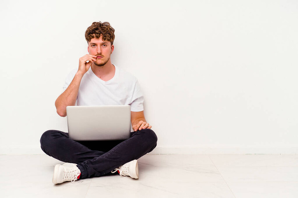 Giovane uomo caucasico seduto sul pavimento tenendo il computer portatile isolato su sfondo bianco con le dita sulle labbra mantenendo un segreto. - Foto, immagini