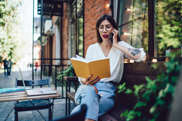Spanish freelancer in classic glasses checking information from memo textbook for discussing project details during international cellphone conversation, millennial woman using mobility connection - Fotografie, Obrázek