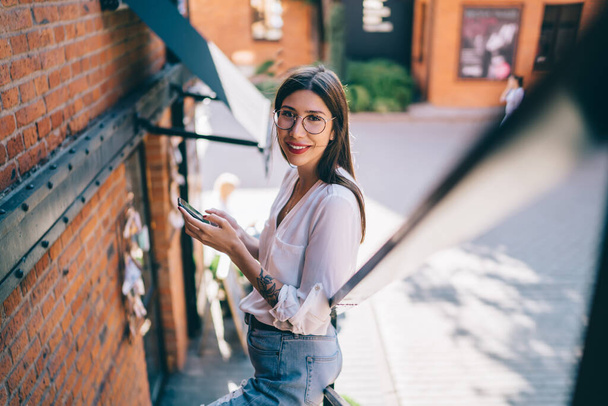 From above of smiling female in trendy eyeglasses with tattoo on hand dressed in white blouse and jeans using mobile phone while standing on stair near building and looking at camera - Foto, imagen