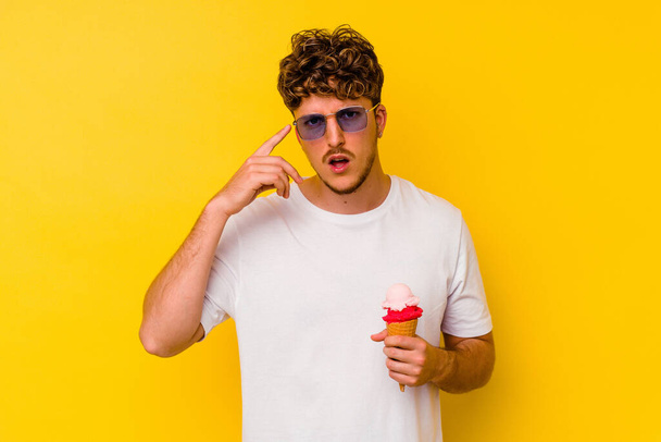 Junger kaukasischer Mann isst ein Eis auf gelbem Hintergrund und zeigt eine enttäuschende Geste mit dem Zeigefinger. - Foto, Bild