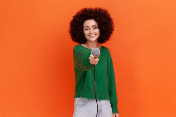 Portrait of satisfied woman with curly hair wearing green casual style sweater standing offering microphone, journalist taking interview. Indoor studio shot isolated on orange background. - Fotografie, Obrázek