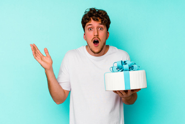 Joven hombre caucásico sosteniendo un pastel aislado sobre fondo azul sorprendido y sorprendido. - Foto, Imagen