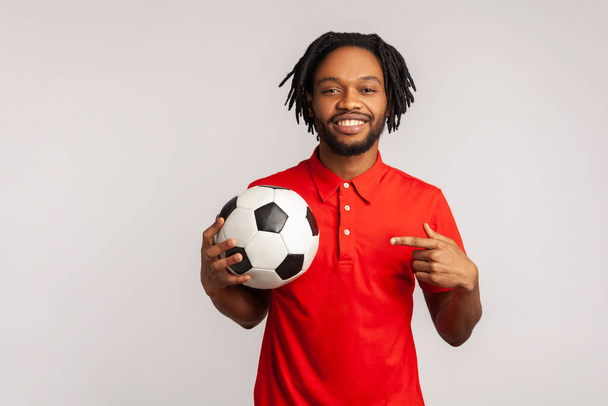 Handsome man with dreadlocks wearing red casual style T-shirt,pointing finger at soccer ball on his hand with smiling positive expression. Indoor studio shot isolated on gray background. - Foto, immagini