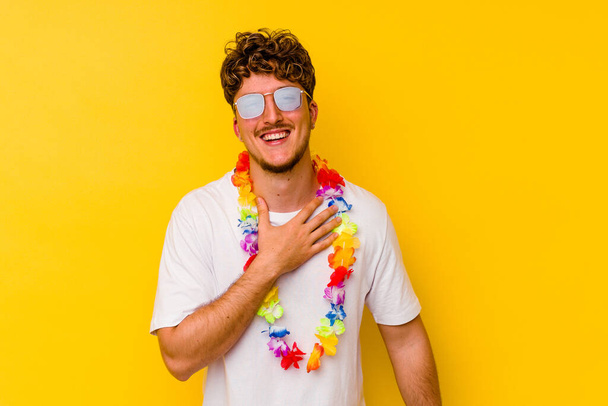 Young caucasian man wearing a Hawaiian party stuff isolated on yellow background laughs out loudly keeping hand on chest. - Valokuva, kuva