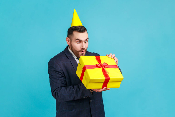 Businessman in official style suit with party cone on head opening gift and looking inside box, unpacking present with expression of funny amazement. Indoor studio shot isolated on blue background. - Foto, Imagen