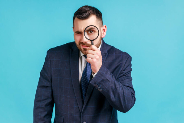 Young adult handsome businessman wearing dark official style suit holding magnifying glass and looking at camera with big zoom eye. Indoor studio shot isolated on blue background. - Fotoğraf, Görsel