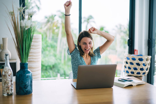 Satisfied young lady in casual dress looking at laptop and raising arms up while receiving news of victory in competition in modern living room - Φωτογραφία, εικόνα