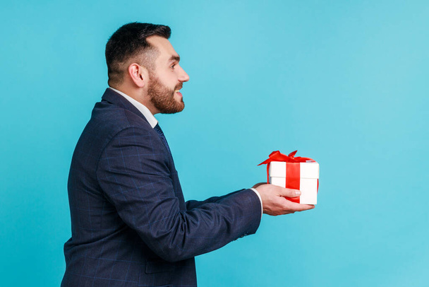Side view of handsome man wearing official style suit giving gift box and smiling broadly, congratulating on holiday and offering present. Indoor studio shot isolated on blue background. - Fotó, kép