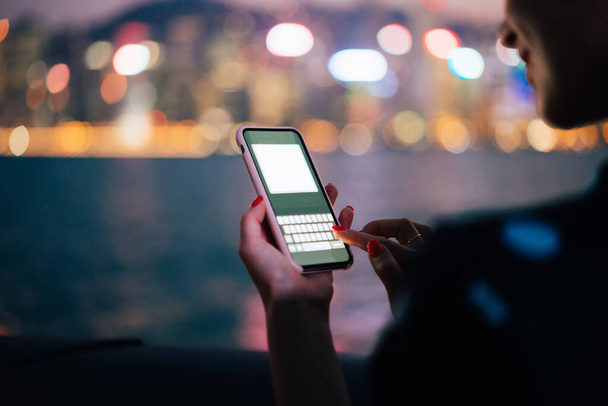 Smartphone moderno em caso rosa com messenger mock up na mão de mulher sorridente irreconhecível cortada no fundo borrado de noite rio e bokeh luzes da cidade - Foto, Imagem