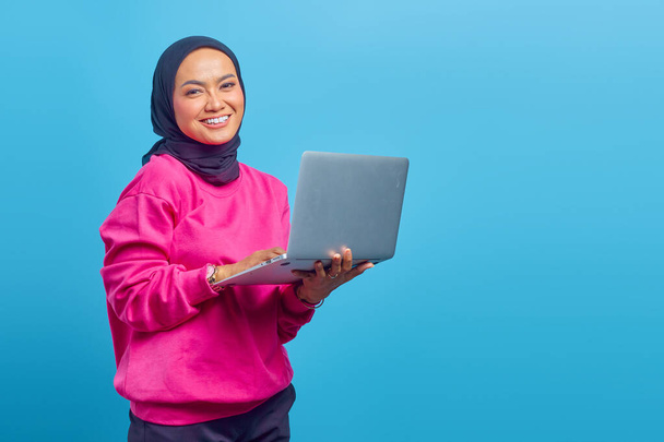 Крупный план азиатской мусульманской девушки, держащей ноутбук в руке на синем фоне - Фото, изображение