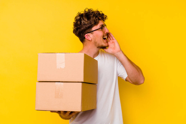 Молодой кавказский мужчина держит картонную коробку, изолированную на желтом фоне, крича и держа ладонь у открытого рта. - Фото, изображение