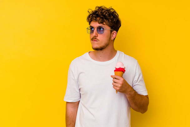 Nuori valkoihoinen mies syö keltaisella taustalla eristettyä jäätelöä unelmoiden tavoitteiden ja päämäärien saavuttamisesta - Valokuva, kuva