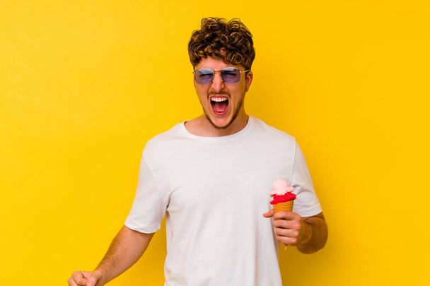 Junger kaukasischer Mann isst ein Eis auf gelbem Hintergrund und schreit sehr wütend und aggressiv. - Foto, Bild