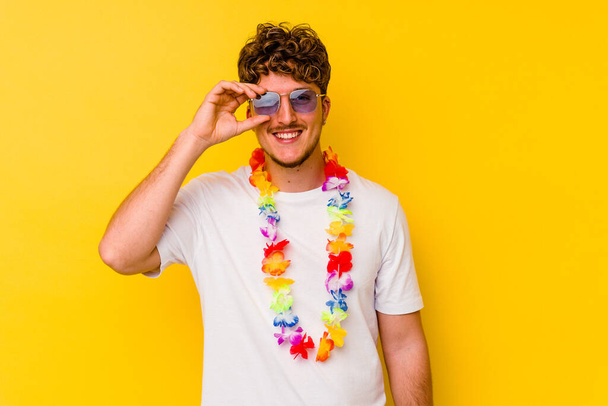 joven caucásico hombre usando un hawaiano fiesta cosas aislado en amarillo fondo excitado mantener ok gesto en ojo. - Foto, imagen
