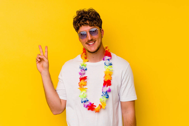 Joven caucásico vestido con una fiesta hawaiana aislado sobre fondo amarillo alegre y despreocupado mostrando un símbolo de paz con los dedos. - Foto, imagen