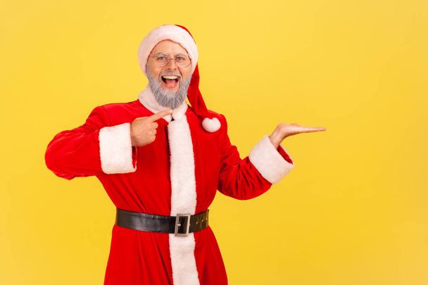 Extremely happy elderly man with gray beard wearing santa claus costume presenting copy space on his palm, looking at camera with open mouth. Indoor studio shot isolated on yellow background. - 写真・画像
