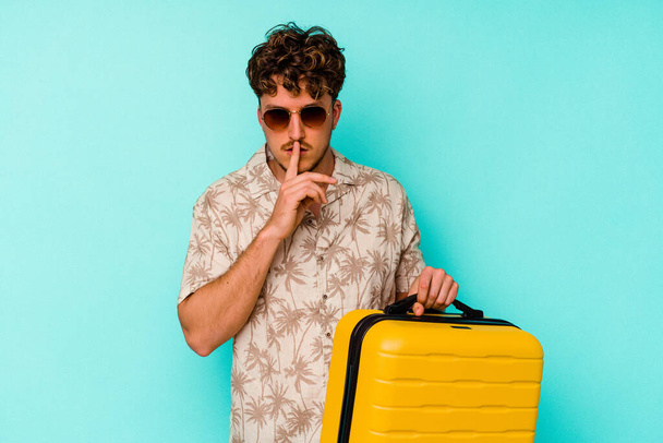 Young traveler man holding a yellow suitcase on blue background keeping a secret or asking for silence. - Фото, зображення