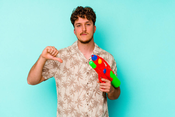 Young caucasian man holding a water gun isolated on blue background showing a dislike gesture, thumbs down. Disagreement concept. - Photo, Image