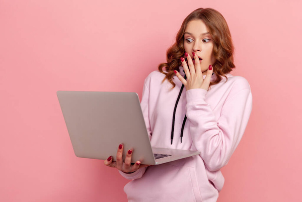 Portrait of shocked curly haired young woman in hoodie covering mouth with palm and looking on laptop display, deadline. Indoor studio shot isolated on pink background - Foto, Imagem