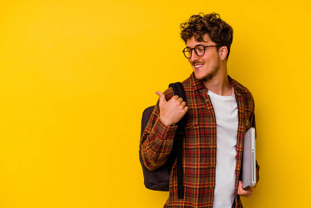 Молодий студент кавказький чоловік тримає ноутбук ізольований на жовтих точках тла з пальцем на великий палець, сміється і безтурботний
. - Фото, зображення