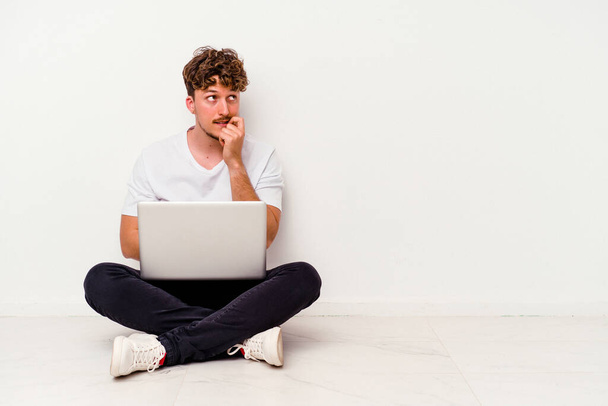 Young caucasian man sitting on the floor holding on laptop isolated on white background relaxed thinking about something looking at a copy space. - Foto, imagen