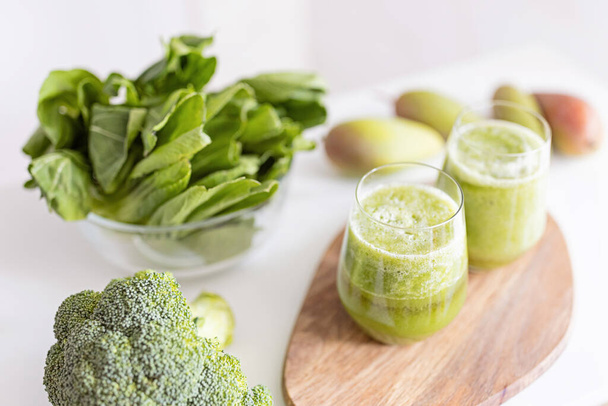 Fresh ingredients for healthy detox cocktail green color. Two glasses with beverage for diet on wooden plate, next lying broccoli and other vegetables and fruits. - 写真・画像
