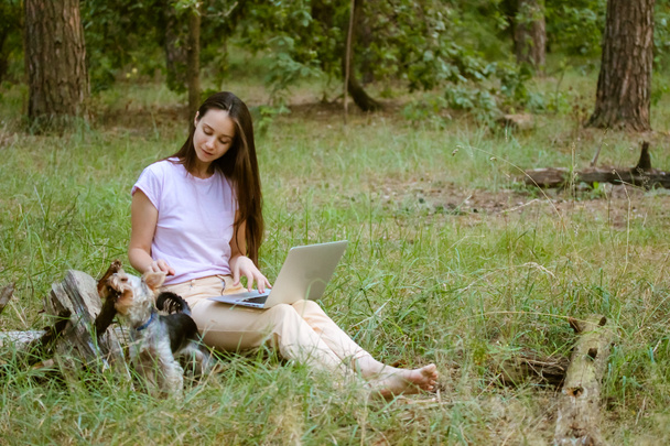 A beautiful young woman, girl with long brown hair sitting in a clearing in green forest with a little dog Yorkshire Terrier, working at laptop. Remote work, study. Student with a computer at nature. - Photo, image