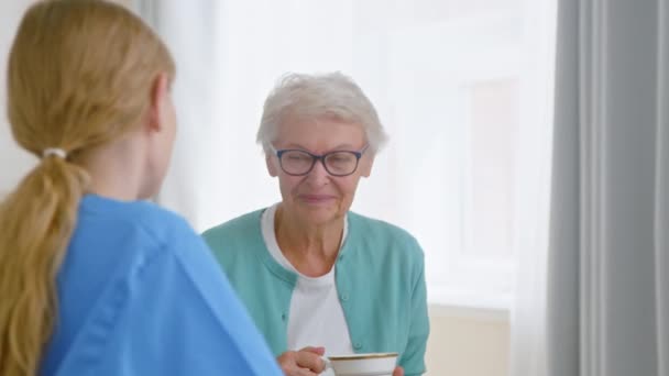 Señora mayor bebe té y habla con la enfermera joven en la habitación - Imágenes, Vídeo