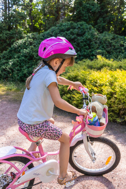 Mała dziewczynka w kasku ochronnym jeździ na dwukołowym różowym rowerze w pięknym parku w słoneczny letni dzień - Zdjęcie, obraz