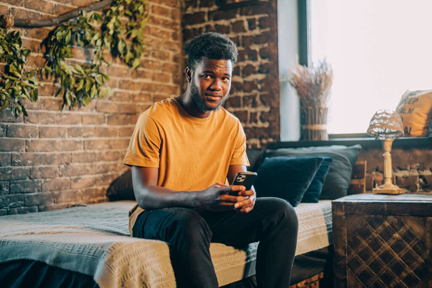 Чорний чоловік пише на своєму мобільному фанаті, сидячи на ліжку вдома
 - Фото, зображення