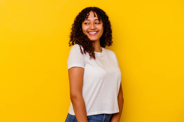 Mladá smíšená rasa žena izolované na žlutém pozadí vypadá stranou s úsměvem, veselý a příjemný. - Fotografie, Obrázek