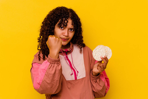 Joven mujer deportiva de raza mixta sosteniendo una torta de arroz aislada sobre fondo amarillo mostrando puño a cámara, expresión facial agresiva. - Foto, Imagen