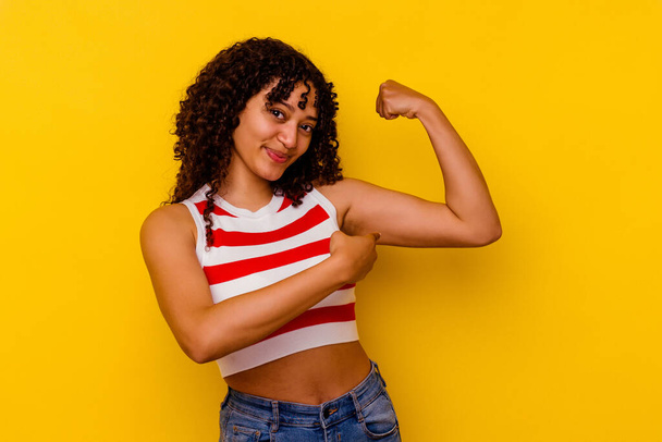 黄色の背景に隔離された若い混合人種の女性は、女性の力の象徴である腕を持つ強さのジェスチャーを示す - 写真・画像