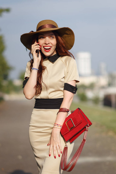 A happy young woman in a wide-brimmed hat is walking in the park in the summer with a bag and talking on the phone - 写真・画像