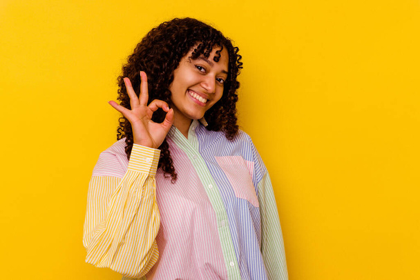 Jonge gemengde ras vrouw geïsoleerd op gele achtergrond knipoogt een oog en houdt een goed gebaar met de hand. - Foto, afbeelding