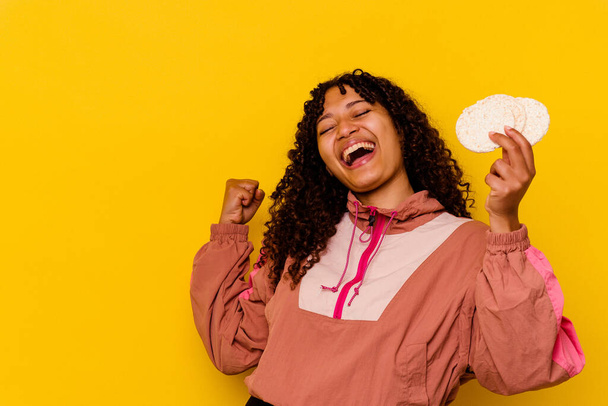 Jonge gemengde ras sport vrouw met een rijst taarten geïsoleerd op gele achtergrond raisen vuist na een overwinning, winnaar concept. - Foto, afbeelding