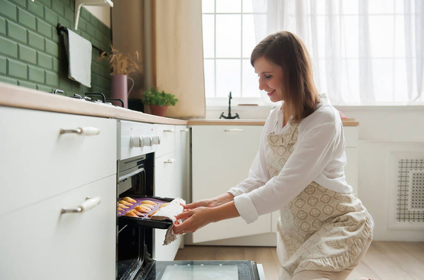 Een jonge blonde vrouw haalt kant-en-klare koekjes uit de oven in de keuken thuis. Een banketbakker. zelfgebakken bakken - Foto, afbeelding