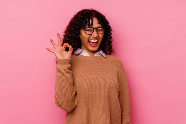 Молодая женщина смешанной расы, изолированная на розовом фоне подмигивает глазом и держит нормальный жест рукой.. - Фото, изображение