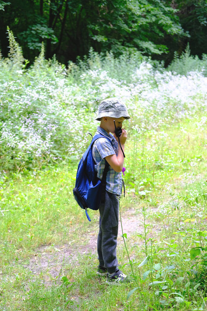 παιδί 7-8 ετών, αγόρι με καπέλο Παναμά, καρό πουκάμισο με κιάλια στο χέρι στέκεται στο δάσος, μιλά σε ένα φορητό ραδιόφωνο, έννοια της πεζοπορίας στη φύση, τον τουρισμό, orienteering, επιβίωση σε άγρια - Φωτογραφία, εικόνα